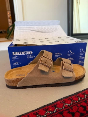 Beige Birkenstock sandaler mocka - Säljer ett par klassiska Birkenstock sandaler i beige mocka med två breda remmar och justerbara metallspännen. Fotbädden är i kork och innersulan är mjuk och bekväm. Yttersulan är i gummi för bra grepp. Perfekta för en avslappnad och stilren look.
