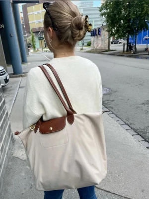 Beige Longchamp Le Pliage handväska - Stilren beige handväska från Longchamp, modell Le Pliage. Väskan har bruna skinnhandtag och lock med guldfärgad knapp. Tillverkad i slitstark syntet med detaljer i skinn. Perfekt storlek för att bära allt du behöver till skolan eller stan. Storlek L💕