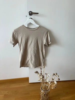 Beige basic t-shirt från H&M - En enkel och stilren beige t-shirt från H&M i mjuk bomull. Modellen är klassisk med rund halsringning och korta ärmar. Perfekt att matcha med jeans eller kjol för en clean look.