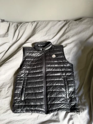 Svart dunväst från Moncler - Snygg svart dunväst från Moncler med klassisk quiltad design och hög krage. Västen har dragkedja framtill, två fickor och Moncler-logga på bröstet. Perfekt för lager-på-lager och streetstyle. Materialet är glansigt och lätt, fyllt med dun.