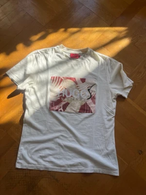 Vit t-shirt från Hugo Boss - Vit t-shirt från Hugo Boss med tryck i rött och beige på bröstet. Klassisk rund hals och korta ärmar. Snygg och enkel design med logga framtill, perfekt till jeans eller shorts.