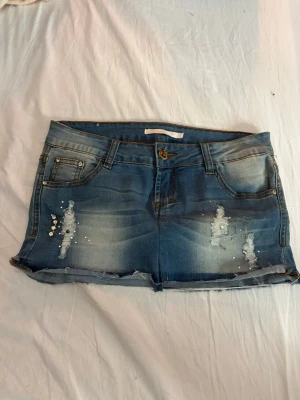 Blå mini jeanskjol med slitningar - Snygg blå mini jeanskjol med slitna detaljer och paljetter både fram och bak. Klassisk femficksmodell med knapp och dragkedja. Perfekta för sommaren och ger en cool, avslappnad vibe.