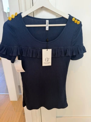 Chiara Forthi Milano topp strl M – helt ny med prislapp - Elegant och feminin mörkblå ribbstickad topp från Chiara Forthi Milano i storlek M. Toppen har snygga guldfärgade knappar på axlarna samt en stilfull volangdetalj över bröstet som ger ett exklusivt och trendigt uttryck.  Helt ny med prislapp kvar – aldrig använd. Perfekt både till vardag och fest, lätt att matcha med jeans, kjol eller kostymbyxor.  ✔ Storlek: M ✔ Färg: Mörkblå ✔ Skick: Helt ny med prislapp ✔ Djur- & rökfritt hem