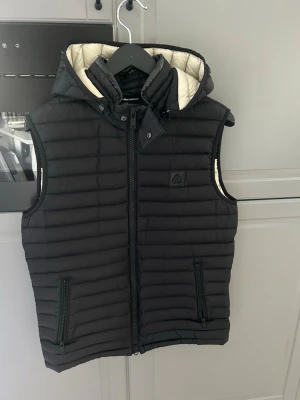 Svart dunväst från Moose Knuckles - Snygg svart dunväst från Moose Knuckles med quiltad design och avtagbar huva. Västen har dragkedja framtill, två fickor med dragkedja och en vit insida som kontrast. Perfekt för lager-på-lager och streetstyle.