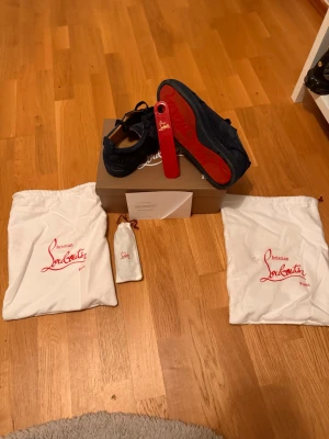 Christian Louboutin svarta sneakers - Snygga svarta sneakers från Christian Louboutin med ikonisk röd sula och broderad logga på sidan. Skorna är i mocka och levereras med originalkartong, skopåse och skohorn. Perfekta för dig som vill sticka ut med exklusiva detaljer och lyxig känsla.