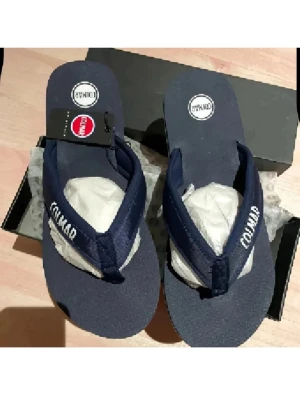 Mörkblå Colmar flip-flops sandaler - Snygga mörkblå flip-flops från Colmar med breda textilremmar och vit logga på remmen. Sulan är platt och har en vit kant runt om. Perfekta för sommaren och lätta att matcha med olika outfits.