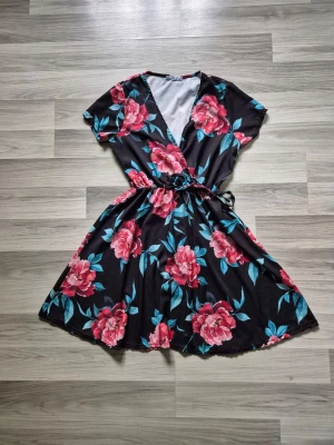 Svart blommig omlottklänning - Svart kortklänning med stora rosa blommor och gröna blad. Omlottmodell med v-ringning, knyt i midjan och korta ärmar. Perfekt för en somrig look med flowy kjol och mjukt material.
