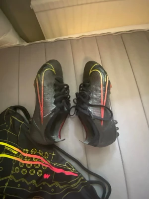 Nike Mercurial Vapor 14 Elite AG - Säljer ett par svarta Nike Mercurial fotbollsskor med gula och röda detaljer. Skorna har snörning och dobbar för spel på gräs. Swoosh-loggan syns tydligt på sidorna och ovandelen är i ett lätt syntetmaterial för bra bollkontroll.