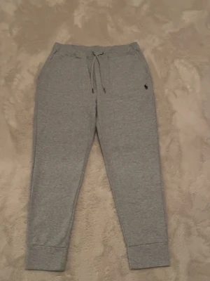 Grå Polo Ralph Lauren byxor - Snygga grå mjukisbyxor från Polo Ralph Lauren med dragsko i midjan och resår vid bensluten. Byxorna har en klassisk passform och är tillverkade i mjukt bomullsmaterial. Liten broderad logga på ena benet ger en stilren touch.