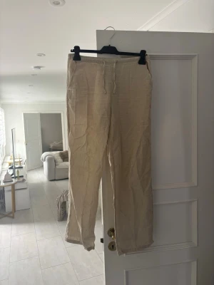 Beiga linnebyxor från Gina Tricot - Säljer ett par beiga linnebyxor från Gina Tricot med avslappnad passform och dragsko i midjan. Byxorna är tillverkade i ett lätt och luftigt linnematerial, perfekta för en chill och stilren look. Passar dig som gillar enkel och bekväm stil.
