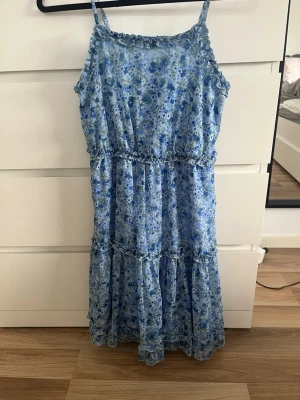 Blommig blå klänning med volang - Söt klänning med blått blommönster och volangdetaljer. Klänningen har smala axelband och ärmlös design, perfekt för varma dagar. Tillverkad i ett lätt och luftigt material som känns skönt mot huden.
