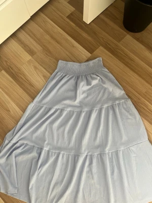 Ljusblå volangkjol med resårmidja - Snygg ljusblå kjol med volanger och bred resår i midjan. Kjolen är lång och har ett luftigt, lätt material som ger ett flowy intryck. Perfekt för dig som gillar en avslappnad men trendig stil.