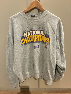 Grå Nike LSU National Champions sweatshirt - Grå sweatshirt från Nike med tryck 'National Champions' i gult och lila samt LSU-logga framtill. Tröja från Nokia Sugar bowl runt 2000 - talet Tröjan har rund halsringning, långa ärmar och avslappnad passform. Perfekt för dig som gillar college-stil och sportiga vibes.