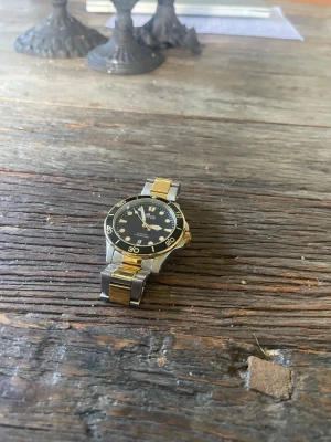 Stilren guldfärgad och silverfärgad klocka - Snygg klocka från Tiga med svart urtavla och guldfärgade detaljer. Armbandet är i metall med både silver- och guldfärgade länkar. Klockan har tydliga siffror och markeringar på boetten, vilket ger en sportig men ändå klassisk look.