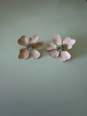 Ljusrosa blomörhängen med strass - Supersöta örhängen formade som blommor i ljusrosa metall med fem hjärtformade kronblad. I mitten sitter en klar strass som ger extra bling. Perfekta för dig som vill ha en feminin och lekfull stil. Stiftet är i metall och örhängena är lätta att bära hela dagen.