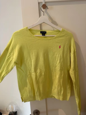 Gul stickad tröja från Ralph Lauren - Snygg gul stickad tröja från Ralph Lauren med rund halsringning och klassisk rosa logga broderad på bröstet. Den har en pytte liten fläck på ena armen. Går säkert bort i tvätten här inte försökt att få bort den 