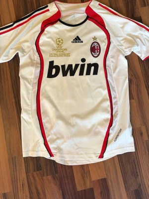 AC Milan Champions Leauge borta tröja 2007 ( KAKA ) - Vit AC Milan matchtröja från Adidas med röda och svarta detaljer, Champions League-finalen 2007 tryckt i guld, klubbmärke och Bwin sponsortryck på bröstet. Tillverkad i lätt och ventilerande material, perfekt för fotbollsträning eller support.