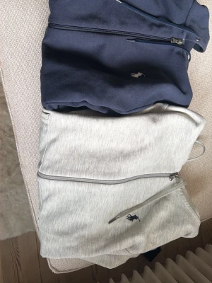 Ralph Lauren ziphoodies i blått & grått - Två snygga ziphoodies från Ralph Lauren, en i marinblått och en i ljusgrått. Båda har dragkedja, huva med dragsko och klassisk Polo-logga på bröstet. Perfekta för en chill och stilren look. Mjukt material och kängurufickor framtill. Helt okej skick lite noppriga. Säljes tillsammans för 500kr, Pris går att diskutera