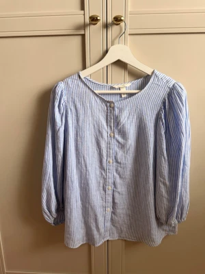 Blårandig linnemixblus från H&M - Snygg blus i blåvitrandigt från H&M, tillverkad i en skön linnemix. Blusen har rund halsringning, knäppning framtill och långa puffärmar med resår vid ärmslut. 