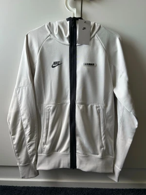 Nike Air Max hoodie med dragkedja - Snygg benvit hoodie från Nike Air Max med svart dragkedja framtill och två fickor. Hoodien har Nike-logga och Air Max-märke på bröstet samt huva. Perfekt för en sportig och avslappnad stil. Aldrig använd prislapp kvar.