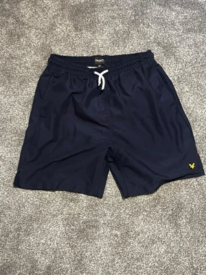 Sprillans nya Mörkblå badbyxor från Lyle & Scott - helt sprillans, nya och oanvända Snygga mörkblå badbyxor från Lyle & Scott med vit dragsko i midjan och klassisk gul logga broderad på vänster ben. byxorna är aldrig använda på grund av en fel beställning. Denna badbyxa passar personer som är mellan 160 cm lång till 180 cm lång.Modellen har sidofickor och elastisk midja för skön passform. Perfekta för stranden eller poolen, och SPECIELLT NU TILL SOMMAREN!. Vid minsta fundering eller fråga var vänligen att meddela mig. Med vänliga hälsningar Eric