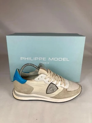 Philippe Model beige sneakers - Storlek 41 men passar 42. Snygga sneakers från Philippe Model i beige och vitt med detaljer i blått och silver. Skorna har mesh och mocka på ovandelen, samt en ikonisk sköldlogga på sidan. Låg modell med vita snören och bekväm sula. Perfekta för en trendig streetstyle-look.