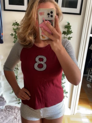Röd baseballtröja med grå ärmar - Säljer en röd baseballinspirerad t-shirt från L.O.G.G. med grå trekvartsärmar och en stor vit siffra 8 på bröstet. Skön och avslappnad passform, perfekt till vardags. Materialet är mjuk bomull.