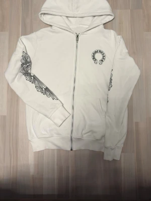 Vit hoodie från Chrome Hearts - Säljer en vit hoodie från Chrome Hearts med dragkedja och huva. Hoodien har svarta grafiska tryck på ena ärmen och ett stort Chrome Hearts-logotyp på bröstet. Tillverkad i mjukt bomullsmaterial och har fickor framtill. Storlek S. Det e inte äkta. 