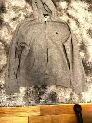 Grå hoodie från Polo Ralph Lauren - Snygg grå hoodie från Polo Ralph Lauren med dragkedja framtill och klassisk huva. Tröjan har det ikoniska lilla Polo-märket broderat på bröstet och är tillverkad i mjukt bomullsmaterial. Perfekt för en chill och avslappnad stil.