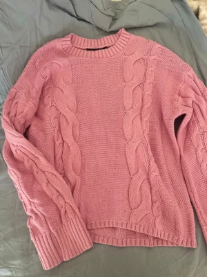 Rosa kabelstickad tröja - Mysig rosa stickad tröja med grova kabelstickade detaljer framtill och på ärmarna. Tröjan har rund halsringning och breda ribbade muddar vid ärmslut och nederkant. Perfekt för kyliga dagar och ger en trendig look.