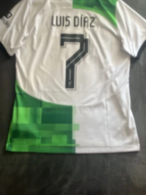 Liverpool Minecraft-kit Luis Díaz #7 - KÖPT I LIVERPOOL! Säljer en Liverpool FC bortatröja från Nike med Luis Díaz och nummer 7 på ryggen. Tröjan är vit med gröna pixelmönster, svarta detaljer och klubbmärke på bröstet. Perfekt för dig som älskar fotboll och vill stötta laget på match eller träning.