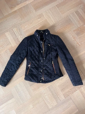 Marinblå quiltad jacka från Zara Woman - Snygg marinblå quiltad jacka från Zara Woman med guldfärgade dragkedjor och röda detaljer. Jackan har hög krage, figurnära passform och två fickor framtill. Perfekt för dig som vill ha en stilren och trendig look till vardags. 