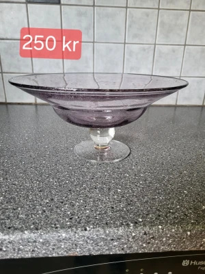 Ljuslila glasskål på fot - Säljer en stilren ljuslila glasskål på fot med bred kant och rundad form. Skålen har en lätt struktur i glaset och passar perfekt som fruktskål eller dekoration. Materialet är glas och färgen är svagt lila.