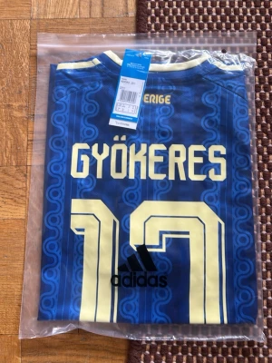 Blå Sverige fotbollströja Adidas - Säljer en blå Sverige fotbollströja från Adidas med Gyökeres och nummer 17 på ryggen. Tröjan har gula detaljer, klassiska Adidas-ränder på axlarna och Sveriges landslagsemblem på bröstet. Tillverkad i lätt och ventilerande material, perfekt för match eller träning.