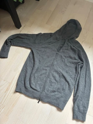 Mörkgrå zip-hoodie gjord i kashmir - Mysig mörkgrå hoodie med dragkedja framtill och två fickor. Tillverkad i kashmir. Klassisk huva och ribbade muddar vid ärmslut och nederkant. Perfekt för en avslappnad stil.