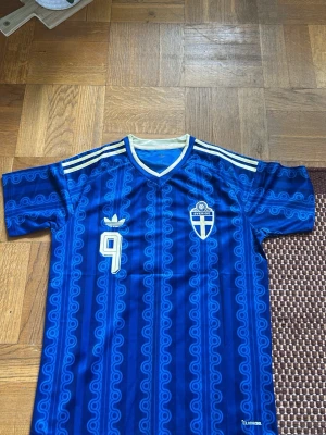 Blå Sverige  fotbollströja Adidas - Retroinspirerad blå fotbollströja från Adidas med Sveriges landslagsemblem och nummer 9 på bröstet. Tröjan har gul/vita detaljer vid krage och axlar samt ett unikt mönster i tyget. Tillverkad i lätt och ventilerande material, perfekt för träning eller match.