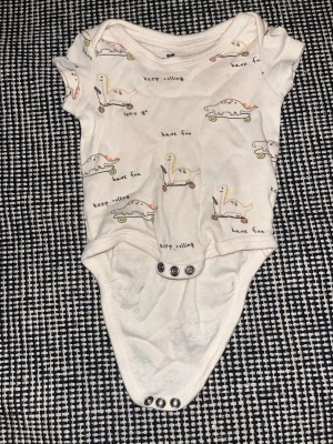 Vit body med dinosaurier från So Cute - Supersöt vit kortärmad body från So Cute med gulligt dinosauriemönster i pastellfärger och små texter som 'have fun' och 'let's go'. Tryckknappar i grenen för enkel påklädning. Perfekt för de allra minsta, 0-3 månader.