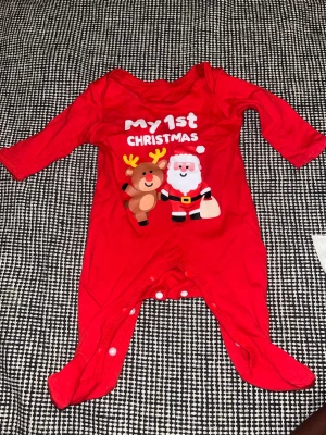 Röd julbody med tomte och ren - Supersöt röd body med tryck av tomte och ren samt texten 'my 1st Christmas'. Långärmad modell med knappar i grenen och täckta fötter. Perfekt för bebisens första jul och tillverkad i mjukt material.