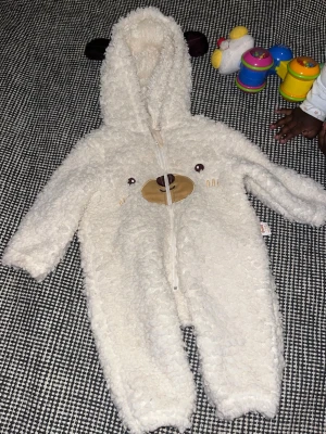 Vit teddyoverall med björnansikte - Supersöt vit teddyoverall med huva och björnöron. Dragkedja framtill och broderat björnansikte på bröstet. Tillverkad i mjukt, fluffigt teddy-material som håller dig varm och bekväm. Perfekt för dig som vill ha en gullig och mysig look.
