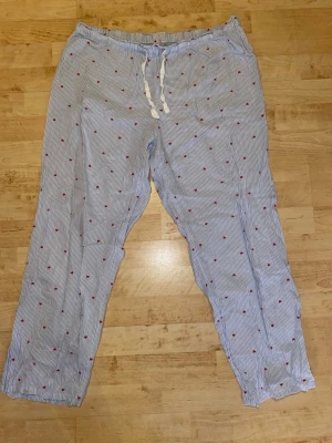 Randiga pyjamasbyxor med små hjärtan - Säljer ett par blå- och vitrandiga pyjamasbyxor med små röda hjärtan. Byxorna har dragsko i midjan och är tillverkade i ett mjukt, tunt tyg som passar perfekt för att chilla hemma. Perfekta för dig som vill ha något bekvämt och gulligt att sova i.