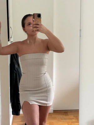 Beige axelbandslös kort klänning - Trendig beige axelbandslös kort klänning med smala vita ränder. Klänningen har en tight passform och en liten slits framtill. Perfekt för dig som gillar en stilren och enkel look. Använd endast 1 gång. 
