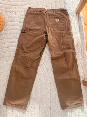 Bruna cargobyxor från Carhartt - Säljer ett par bruna cargobyxor från Carhartt med raka ben och flera praktiska fickor. Byxorna har klassisk Carhartt-logga på bakfickan och är tillverkade i slitstark bomullscanvas. Perfekta för dig som gillar streetwear och funktionella detaljer. Storlek 34 x 32