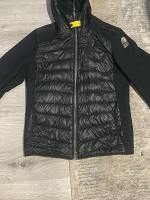 Svart cardigan från Parajumpers - Säljer en svart cardigan från Parajumpers med quiltad front, stickade ärmar och huva. Jackan har gul detalj vid halsen, bröstficka med dragkedja och klassisk Parajumpers-logga på ärmen. Perfekt för kalla dagar och riktigt snygg streetstil.
