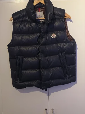 Svart dunväst från Moncler - Snygg svart dunväst från Moncler med hög krage och två dragkedjefickor framtill. Västen har Monclers klassiska logga på bröstet och är tillverkad i glansigt material med dunfyllning. Perfekt för lager på lager och streetwear-stil.