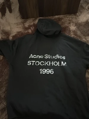 Svart hoodie Acne Studios Stockholm - Cool svart hoodie från Acne Studios med stor vit text på ryggen: 'Acne Studios STOCKHOLM 1996'. Klassisk passform med huva och ribbade muddar. Perfekt för dig som gillar streetwear och vill ha något från ett ikoniskt svenskt märke.