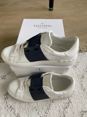Valentino Garavani vita sneakers - Valentino Garavani sneakers i mycket fint skick. Använda färre än 10 gånger, inga fel eller skador. Vita lädersneakers med mörkblå rem och nitdetaljer vid hälen. Stilrena, bekväma och tidlösa.