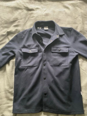 Mörkblå overshirt från Selected Homme - Stilren mörkblå overshirt från Selected Homme med två bröstfickor med lock och knappar. Skjortan har klassisk krage och knäppning framtill. Perfekt lager-på-lager-plagg i mjukt material.