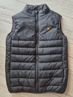 Svart dunväst från Lyle & Scott - Snygg svart dunväst från Lyle & Scott med gul logga på bröstet. Västen har hög krage, dragkedja framtill och quiltad design. Perfekt att slänga över en hoodie eller tröja för extra värme. Klassisk och stilren look som funkar till det mesta.