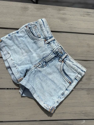Ljusblå denimshorts med rå kant - Snygga ljusblå denimshorts med råa kanter och klassisk femficksdesign. Shortsen har bälteshällor, knapp och dragkedja framtill samt slitna detaljer för en avslappnad look. Perfekta för varma dagar och enkel att matcha med olika toppar. Shortsen har fransiga kanter och är Low waist😊