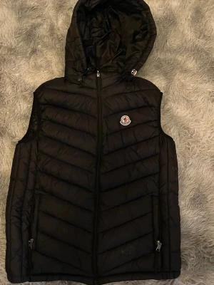 Svart dunväst med huva från Moncler - Snygg svart dunväst från Moncler med huva och dragkedja framtill. Västen har två fickor med dragkedja och Moncler-logga på bröstet. Tillverkad i glansigt material och har ett modernt, vadderat mönster. Perfekt för lager-på-lager och streetstyle. (EJ ÄKTA) Kan sänka vid snabbaffär